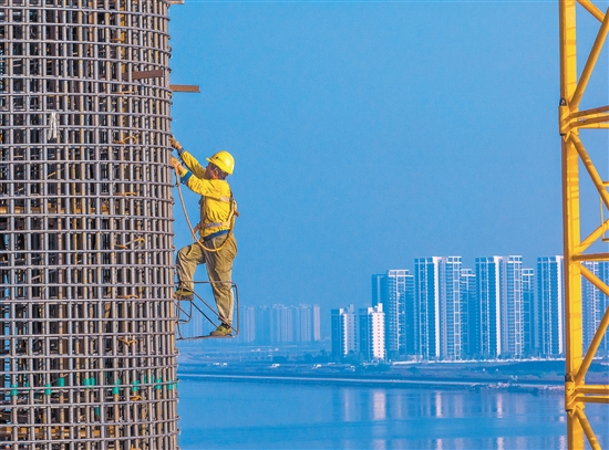 奮戰(zhàn)在百米索塔之上的建設(shè)者身影，與遠處的高樓大廈遙呼相應(yīng)。